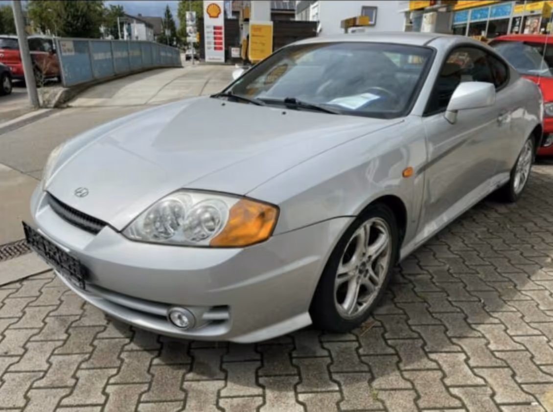 Silver Hyundai Coupé GK in stock condition, front three-quarter view at a petrol station