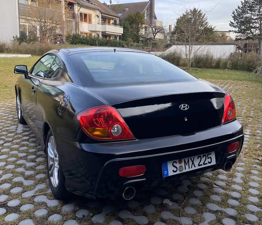 The same Hyundai Coupé GK after the conversion, painted black, rear three-quarter view showing the body kit and dual exhaust